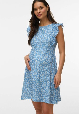 Gravid kvinde med langt mørkt hår iført en blå blomsterprintet, ærmeløs kjole, der blidt holder sin mave, stående foran en hvid baggrund.