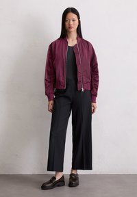 Woman wearing a burgundy bomber jacket, black wide-leg pants, black top, and dark brown loafers standing against a plain light wall.