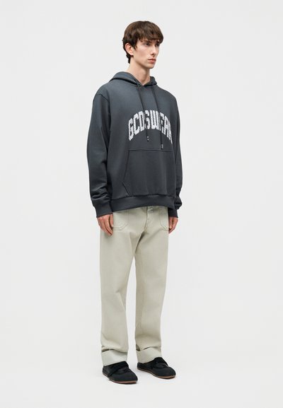 Young man standing, wearing a dark gray hoodie with white text and light beige pants with black shoes on a plain white background.