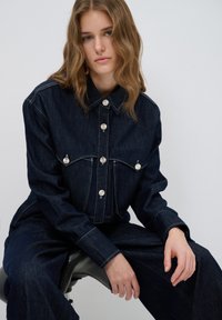 Tumma farkkutakki, jossa on kaulus, etutaskut ja hopeanväriset napit. Kangas on sileä ja siinä on kontrastisia valkoisia ompeleita.