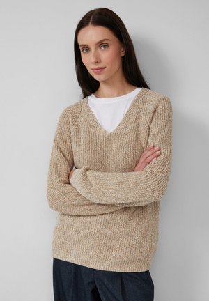 Frau mit langen, dunklen Haaren, die einen beigen, gestrickten V-Ausschnitt-Pullover über einem weißen Hemd trägt, mit übergeschlagenen Armen, stehend vor einem schlichten, hellen Hintergrund.