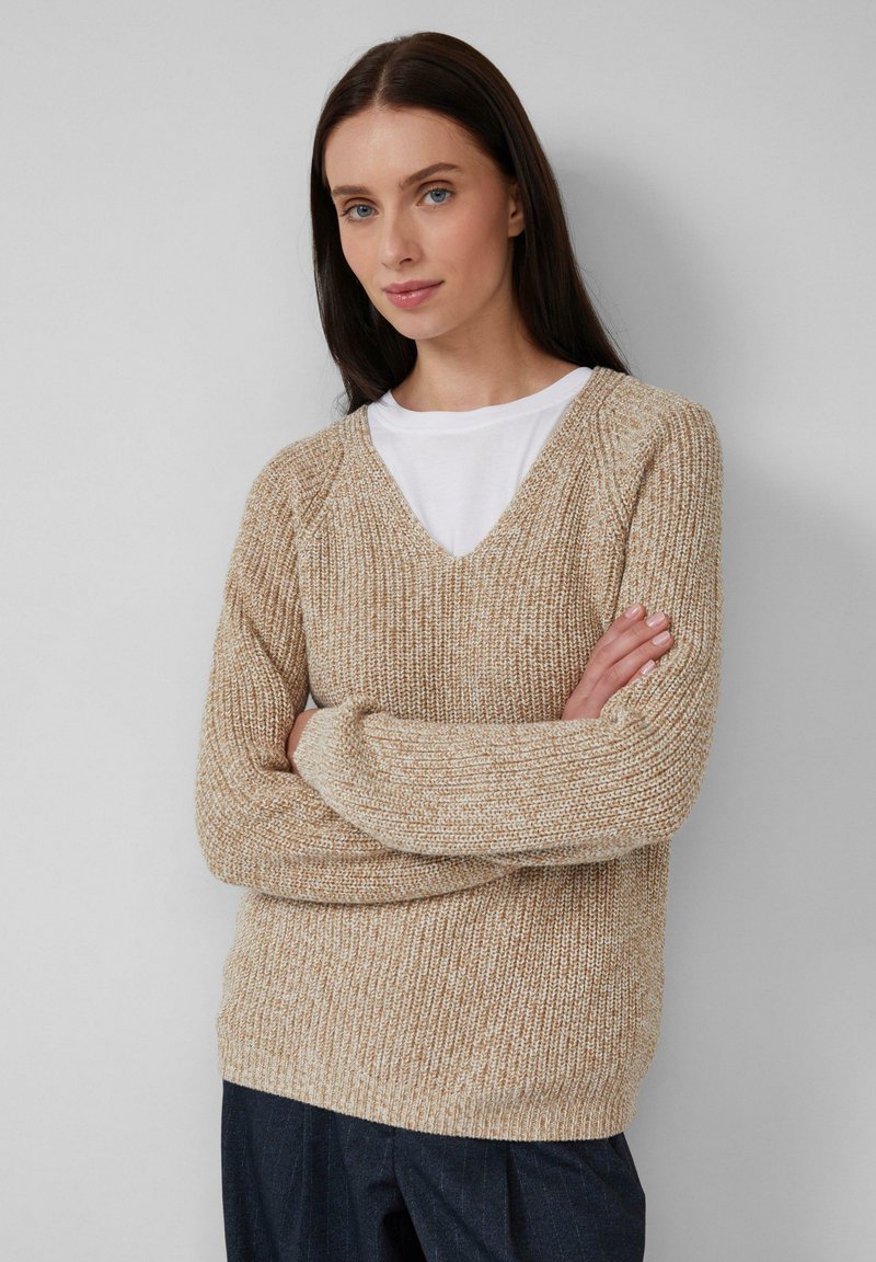 Frau mit langen, dunklen Haaren, die einen beigen, gestrickten V-Ausschnitt-Pullover über einem weißen Hemd trägt, mit übergeschlagenen Armen, stehend vor einem schlichten, hellen Hintergrund.