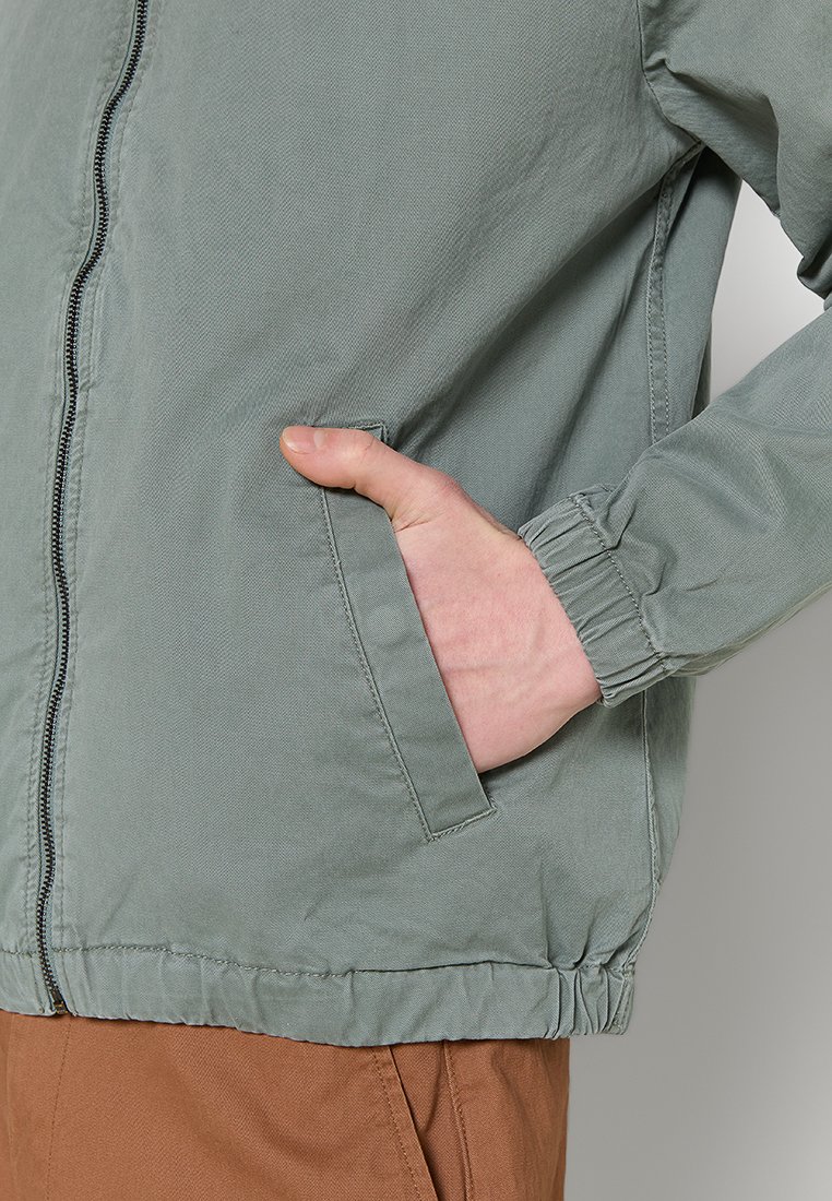Helle grüne Jacke aus strukturiertem Stoff, mit Reißverschluss vorne, seitlichen Taschen und elastischen Bündchen.