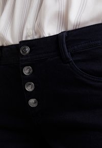 Tmavě modré džíny mají pětibuttonové zapínání s kovovými detaily, ploché přední kapsy a hladkou texturu, zobrazené vedle pruhované látky.