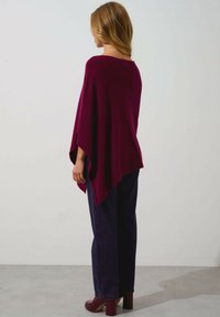 Poncho bordeaux con una vestibilità morbida, realizzato in tessuto morbido, caratterizzato da un ampio scollo e orlo asimmetrico abbinato a jeans a gamba larga di colore blu scuro.
