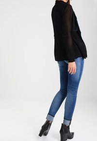 Svart stickad cardigan med strukturerad design, långa ärmar och öppen front, kombinerad med blå denimjeans och svarta höga ankelstövlar.