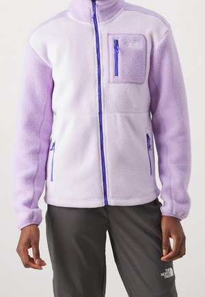 Personne portant une veste polaire lavande et blanche avec des fermetures éclair bleues et un pantalon gris foncé, debout devant un fond uni.