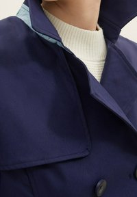 Námořnicky modrý kabát s vysokým límcem, hladkou texturou a dvouřadým zapínáním. Lehčí látka pod límcem dodává kontrast.