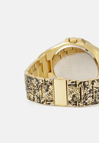 Gold-tone wristwatch with a black patterned band featuring intricate designs. Includes a silver-toned caseback and push-button clasp.