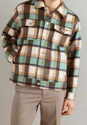 Persona con una chaqueta de cuadros en verde, marrón, beige y blanco con botones y bolsillos, combinada con pantalones marrón claro.