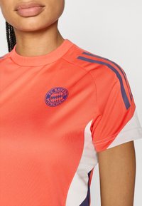 Φωτεινό κοραλλί αθλητικό μπλουζάκι με ναυτικά στοιχεία, με το λογότυπο της FC Bayern Munich στο στήθος. Κατασκευασμένο από μαλακό, ελαστικό ύφασμα.