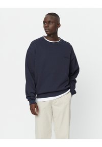 Felpa girocollo bleu navy realizzata in morbido tessuto, con una vestibilità comoda e un logo discreto sul petto, abbinata a pantaloni beige chiaro.