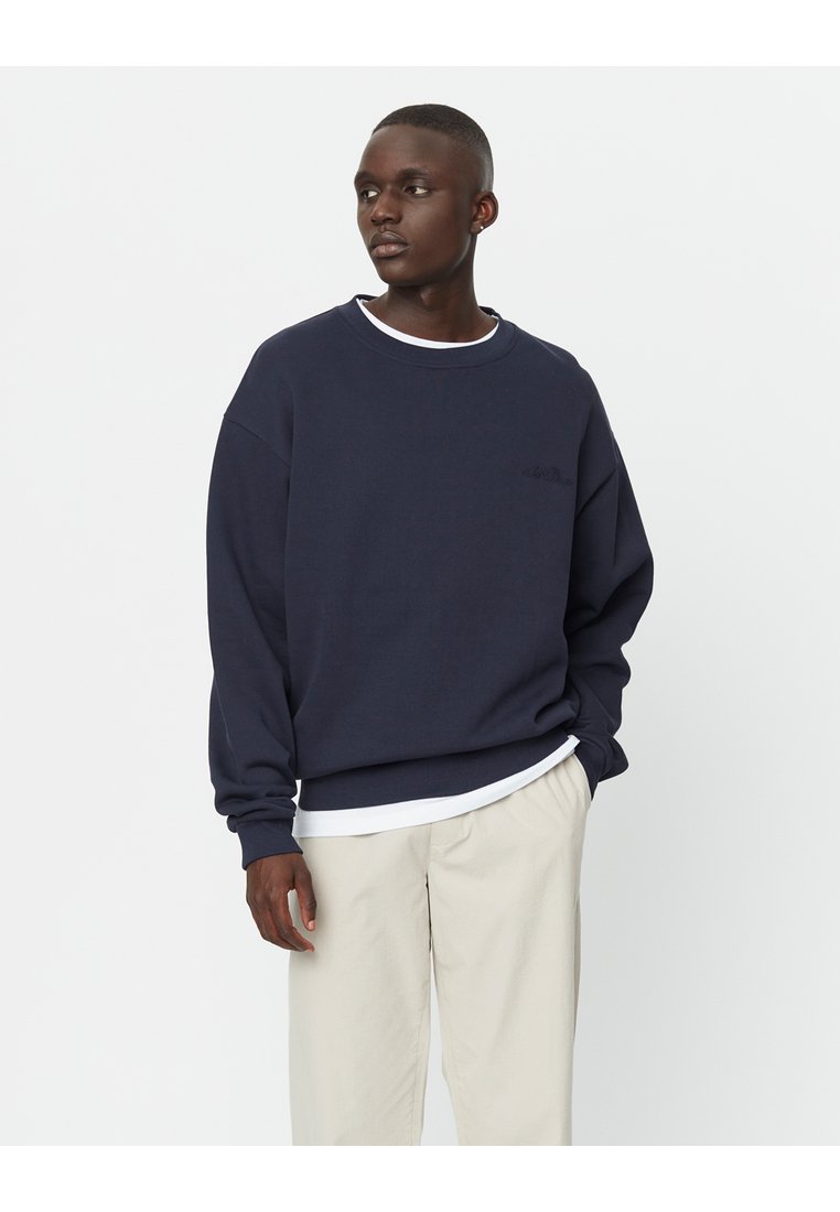Felpa girocollo bleu navy realizzata in morbido tessuto, con una vestibilità comoda e un logo discreto sul petto, abbinata a pantaloni beige chiaro.