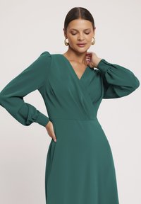 Zielona sukienka z długim rękawem, uszyta z gładkiego materiału, z dekoltem w kształcie litery V, marszczonymi rękawami oraz dopasowaną talią i zwiewną częścią spódnicy.