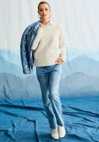 Vrouw in een beige gewatteerde sweater, lichtblauwe spijkerbroek en witte laarzen, die een blauwe pufferjack over haar schouder houdt met een blauwe bergachtergrond.