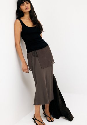 Pikkade tumedate juustega naine, kes kannab musta tanktoppi, pruuni asümmeetrilist seelikut ja musti rihmikuga kontsakingi, toetudes valgele seinale.