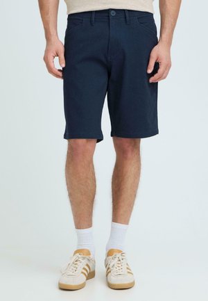 Homme portant un short bleu marine, des chaussettes blanches et des baskets beiges, debout devant un fond gris clair.