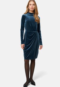 Samtige türkisfarbene Kleid mit langen Ärmeln, hohem Ausschnitt und verdrehten Details an der Taille. Kombiniert mit schwarzen Strumpfhosen und spitzen Ballerinas.