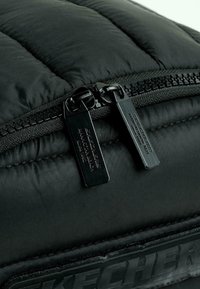 Material acolchado negro con textura acolchada, que presenta dos cremalleras negras y acentos metálicos, además de un logo moldeado en el costado.