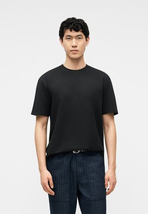 Joven de pie mirando al frente, vistiendo una camiseta negra lisa de manga corta y pantalones azul oscuro con rayas y bolsillos parche.