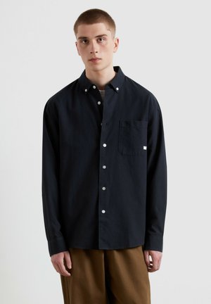 Giovane uomo con capelli corti che indossa una camicia nera abbottonata e pantaloni marroni, in piedi davanti a uno sfondo chiaro e uniforme.