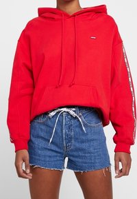 Red hooded sweatshirt with a front pocket and drawstrings, paired with blue denim shorts featuring frayed edges and a patterned waistband.