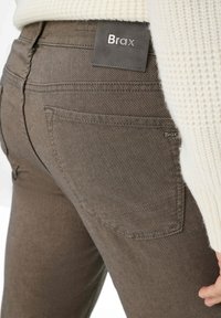 I jeans in denim stretch marrone presentano un motivo a tessitura strutturata, una tasca posteriore visibile e un'etichetta con il logo Brax sulla vita.