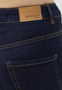 Jeans in denim scuro con cuciture arancioni, dotati di una etichetta in pelle con la scritta "perfect jeans" e un design per la tasca posteriore. Texture liscia.