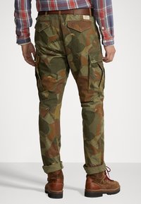 Pantalones cargo de camuflaje verde con detalles bordados, ajuste entallado, puños enrollados y bolsillos traseros, combinados con botas marrones de cordones.