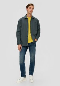Graue Button-Down-Jacke, darunter ein gelbes T-Shirt, blaue Jeans und weiße Sneakers. Die Jacke hat eine Brusttasche und eine lässige Passform.