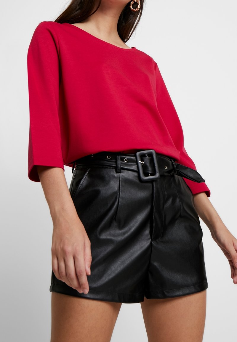 Femme portant un haut rouge à manches trois-quarts et un short noir en cuir synthétique taille haute avec une large ceinture et une grande boucle.