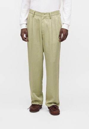 Mand iført lyse beige plisserede bukser, en hvid langærmet sweater og brune snøresko, stående foran en hvid baggrund.