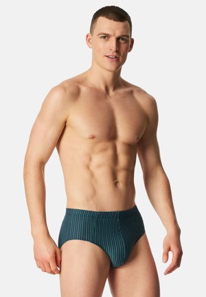 Man zonder shirt die staat, draagt donkerteal slip met dunne verticale witte strepen tegen een effen lichte achtergrond.