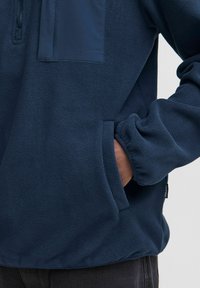 Pullover in pile blu navy con colletto zip, polsini elasticizzati e tasca a toppa; texture morbida con un dettaglio di pannello liscio sul petto.