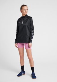 Zwarte langemouwtop met een kwart rits en een grijs chevronpatroon op de mouwen, gecombineerd met roze shorts en blauwe sportschoenen.