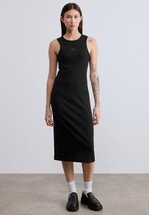 Abito midi nero a coste senza maniche con scollo rotondo, silhouette aderente e dettaglio logo, abbinato a scarpe slip-on nere e calzini bianchi.