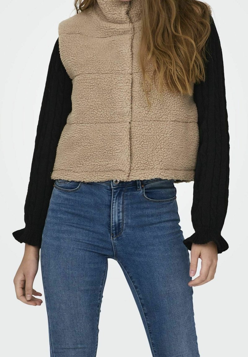 Femme portant un gilet sherpa beige par-dessus un pull noir à manches longues et un jean taille haute bleu, debout devant un fond blanc.