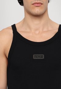Čierny rebrovaný top s okrúhlym výstrihom, na prednej strane má štvorcový čierny štítok s nápisom "VERSACEJEANS COUTURE".