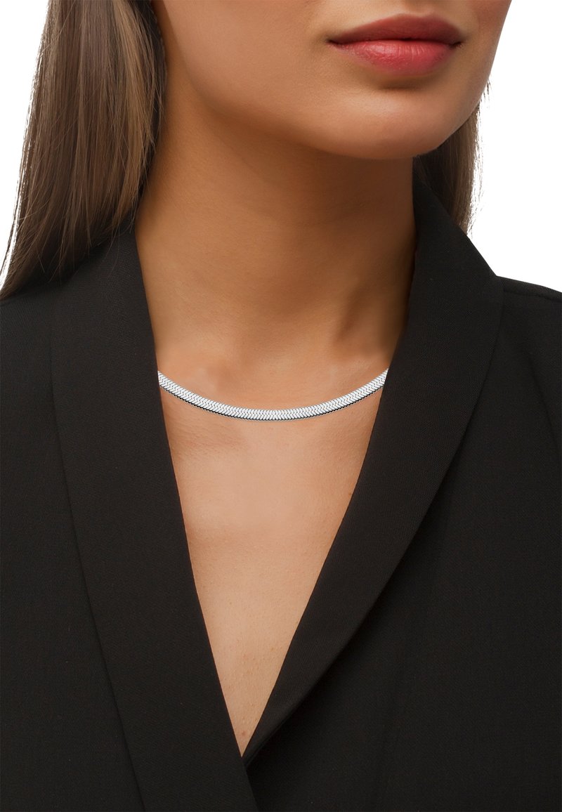 Collier ras du cou en métal argenté avec un design lisse et plat, mettant en valeur une surface réfléchissante sur un fond de tenue sombre.