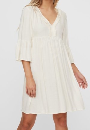 Løs hvid kjole med V-udskæring, dekorativ kant og tre-kvart lange klokkeærmer, båret af en stående person.