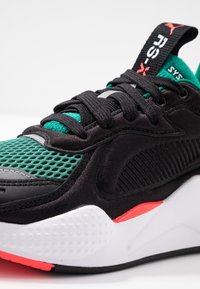 Svart atletisk sneaker med gröna nät- och läderdetaljer, vit sula, röda detaljer och svarta snören. Texturerade ytelement ingår.