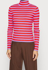Tätt ribbad polotröja med horisontella röda och lila ränder, i kombination med lösa bruna byxor.