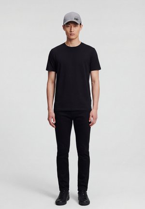 Mannelijk model staand, met een grijs baseballpetje, zwart T-shirt met korte mouwen, zwarte broek en zwarte sneakers op een effen grijze achtergrond.