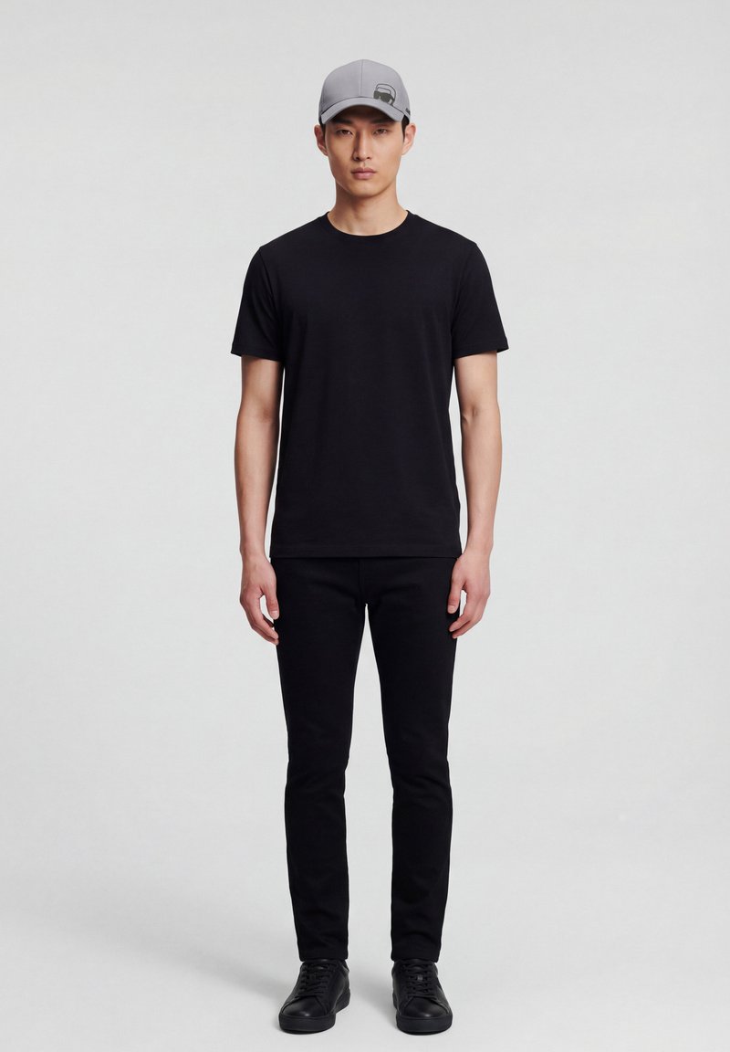 Modèle masculin debout, portant une casquette de baseball grise, un t-shirt noir à manches courtes, un pantalon noir et des baskets noires sur un fond gris uni.