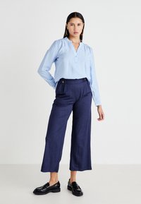 Camisa de botones a rayas azul claro con mangas abullonadas, combinada con pantalones de pierna ancha azul marino con detalles dorados y mocasines negros.