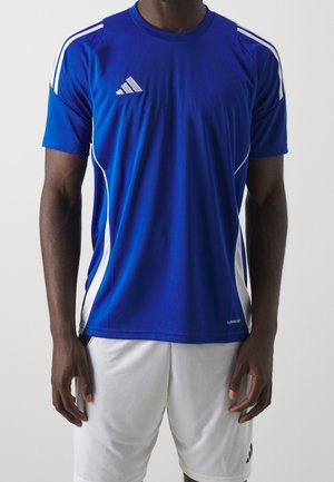 Homme portant un maillot de sport bleu Adidas avec des bandes blanches et un short de sport blanc, debout devant un fond uni.