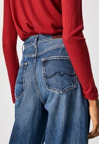 Person som bär högmidjade blå denimjeans med sydda bakfickor och en instoppad långärmad röd ribbad topp.