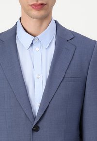 Helles blaues Sakko aus feinem Stoff, mit einem einzelnen schwarzen Knopf und einem passenden hellblauen Hemd darunter.