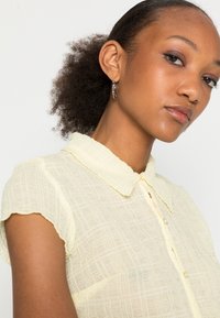 Blusa amarela transparente com gola, padrão de grelha texturizada, mangas curtas e botões semelhantes a pérolas na frente.