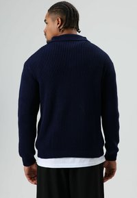 Man met ingevlochten haar draagt een marineblauwe geribbelde trui over een wit overhemd, kijkt weg tegen een effen lichtgrijze achtergrond.